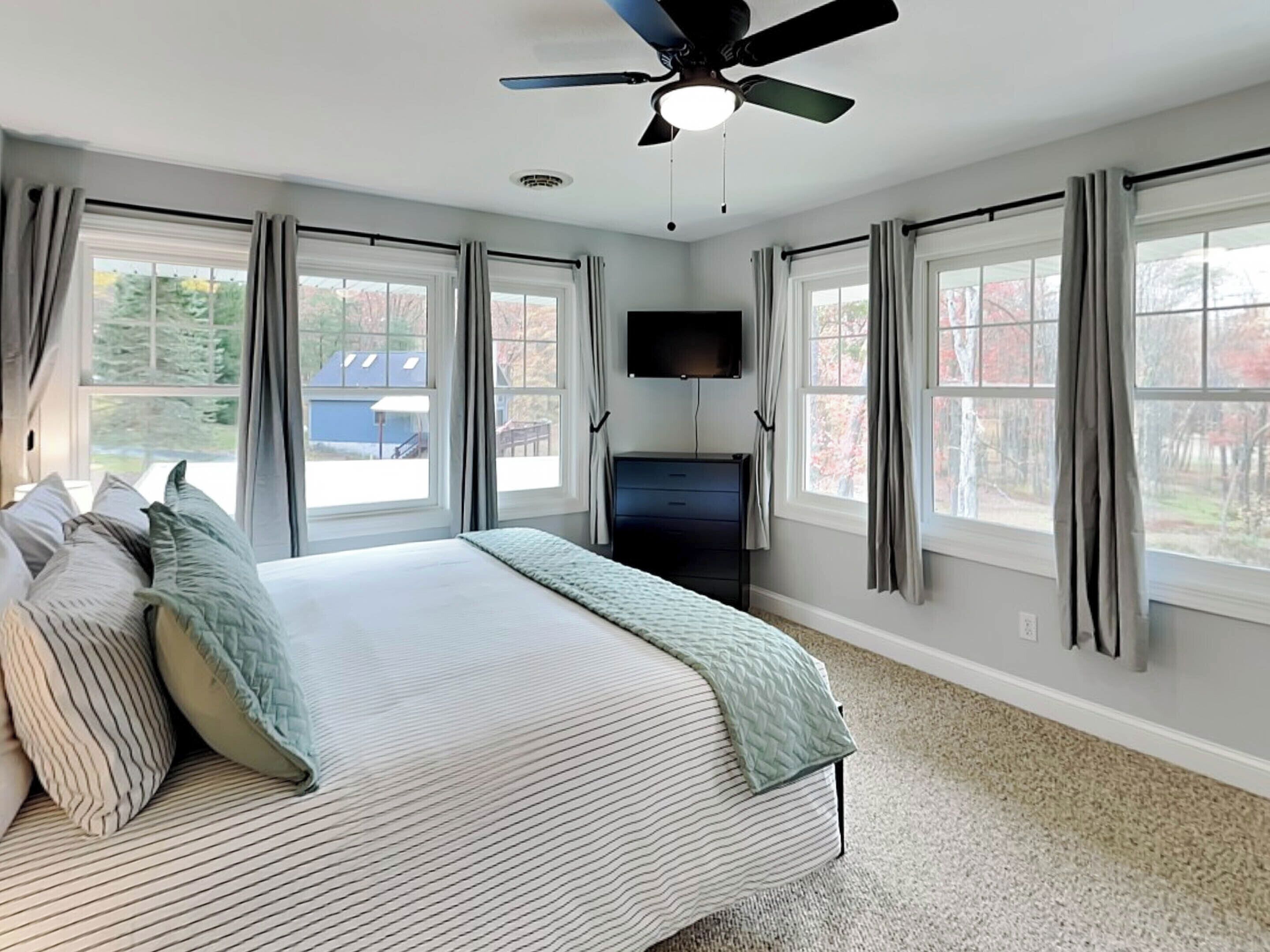 A bedroom with a bed, a ceiling fan, a TV, and large windows with curtains.