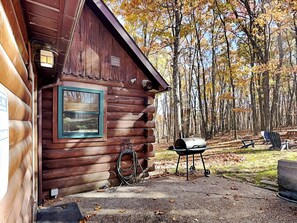 Una cabaña de troncos con una ventana, una parrilla de barbacoa y una silla afuera.