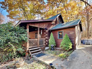 Une cabane rustique en rondins de bois, dotée d’un porche, de deux arbres à feuilles persistantes et d’une pancarte de bienvenue.