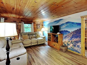 A cozy living room with a wooden ceiling, a beige sofa, a television on a wooden stand, and a scenic mountain mural on the wall.