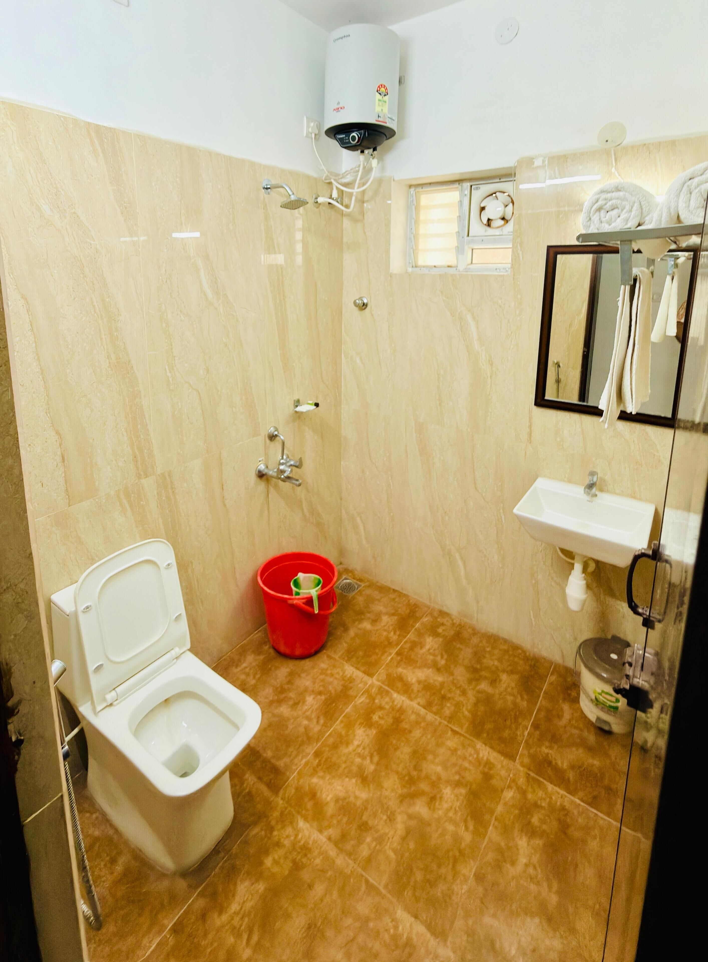 A bathroom with a toilet, a red bucket, a shower, a sink, and a mirror.