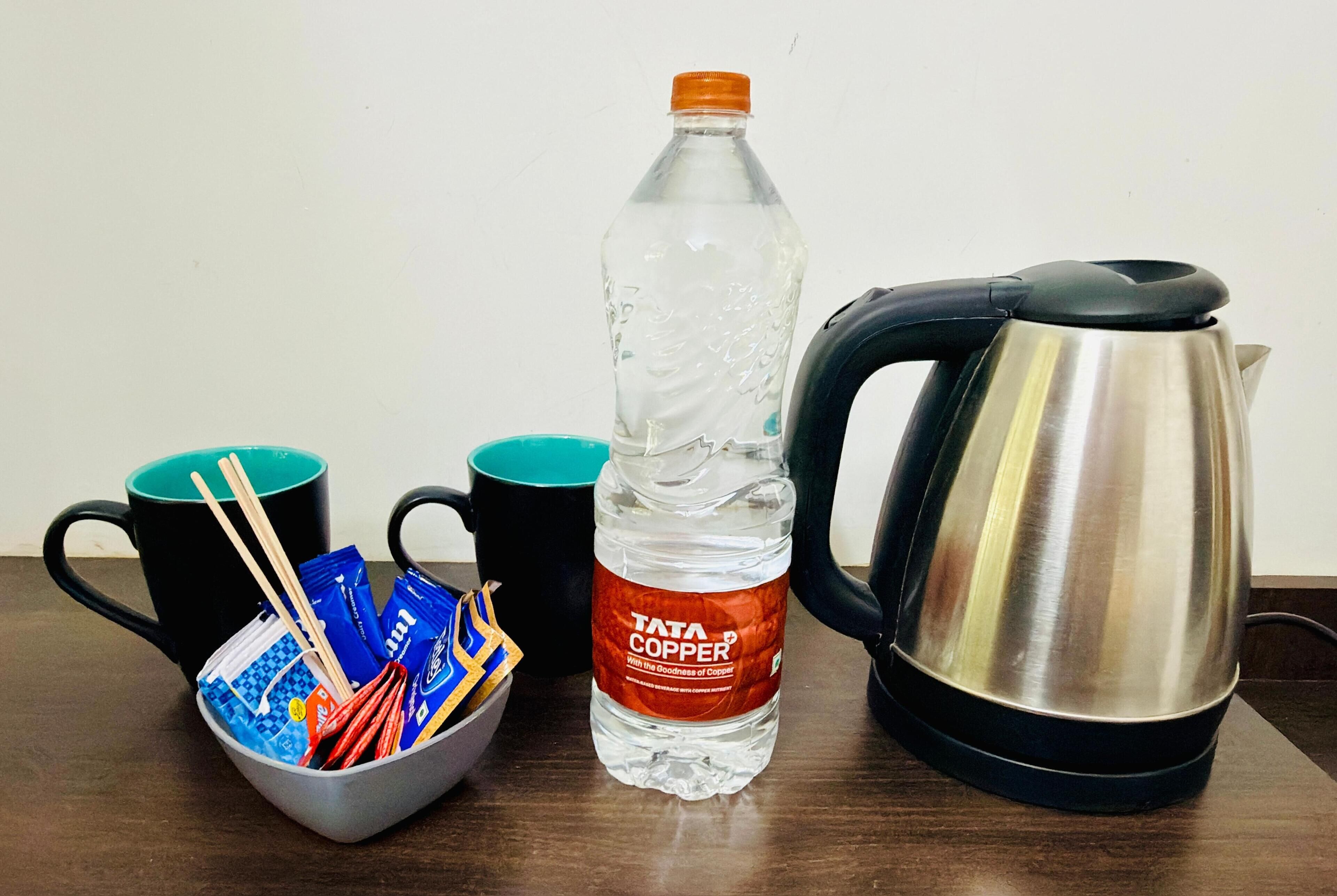 Una mesa con un hervidor negro, una botella transparente de agua, dos tazas color turquesa y un bol con aperitivos y palillos.