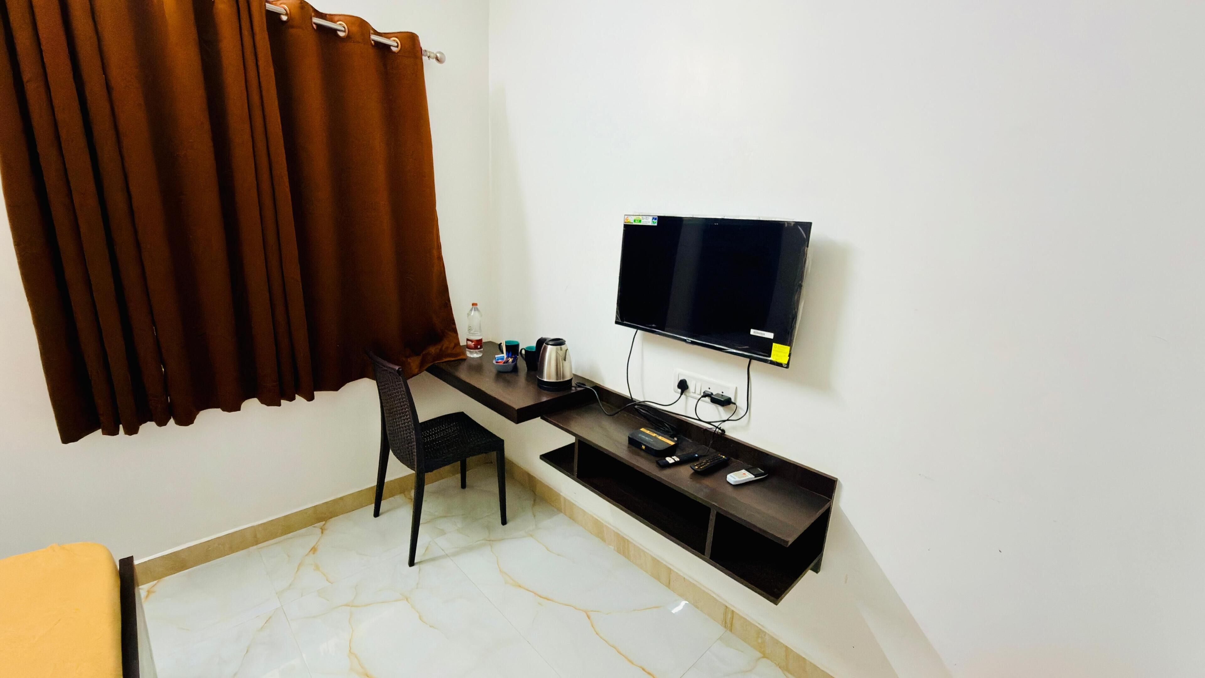 A small, modern room with a flat-screen TV mounted on the wall, a desk with a chair, and a window with brown curtains.