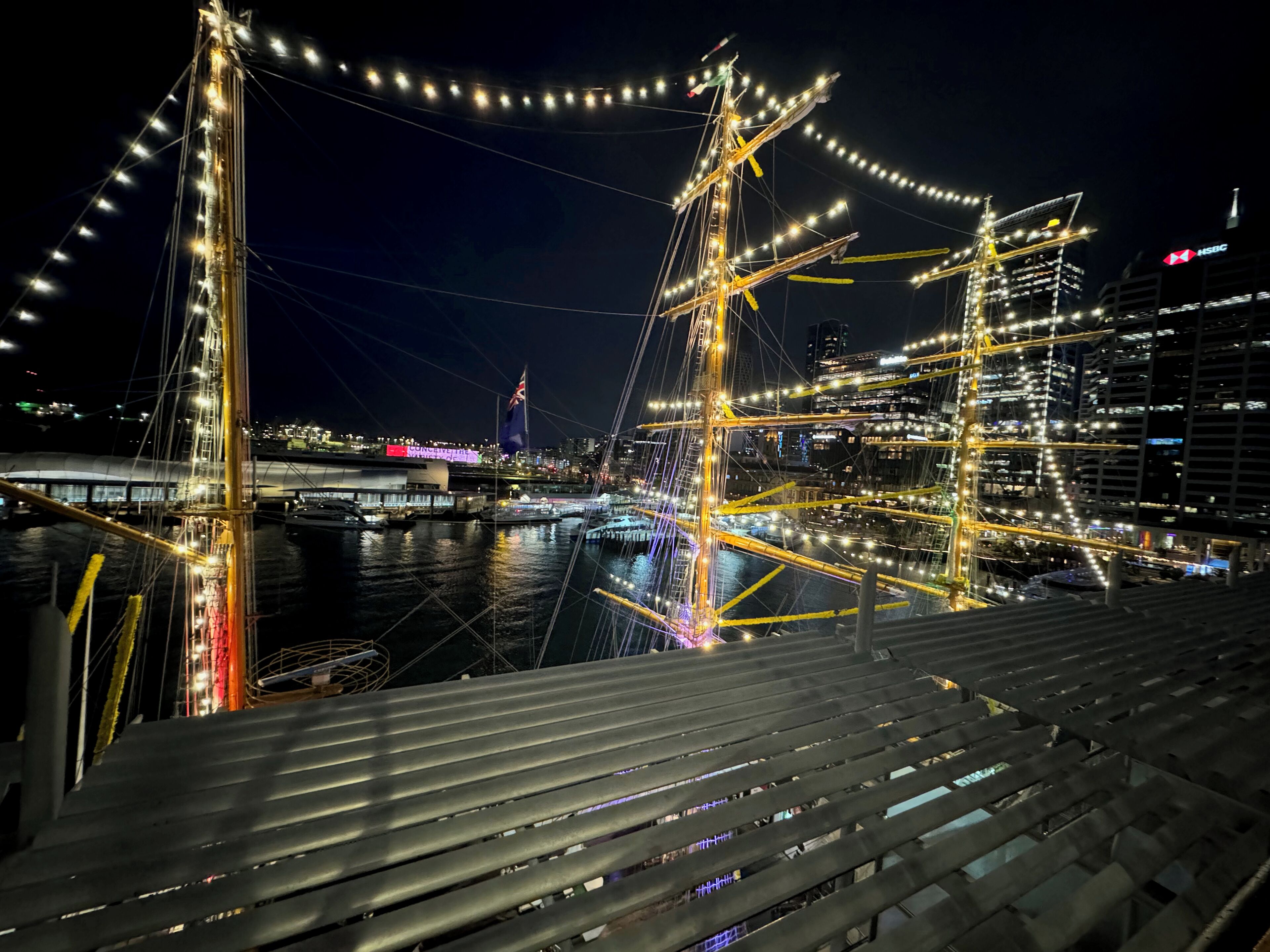 Un puente grande e iluminado con múltiples cables y una bandera, con un paisaje urbano que incluye edificios y un estadio.
