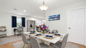 A dining area with a white table set for four, grey chairs with gold legs, a chandelier, a white door, and a window with blue curtains. - Lake George Village -1 Block to Beach, Lake, Town. Newly Remodeled and Spacious. (Lake George)