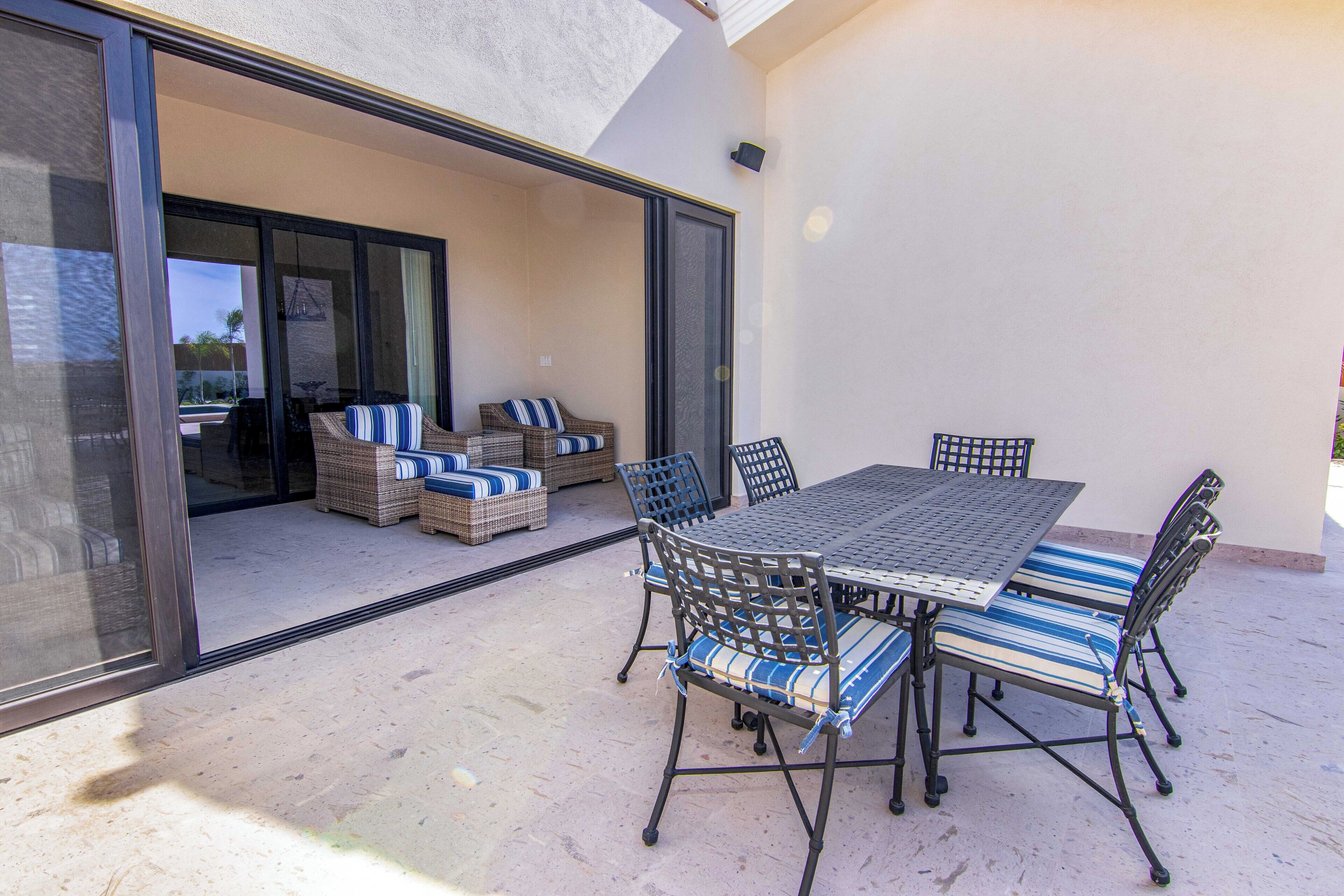 Eine Terrasse im Freien mit Tisch und Stühlen, eine Rattan-Couch mit gestreiften Kissen und eine Glastür zu einem überdachten Bereich.