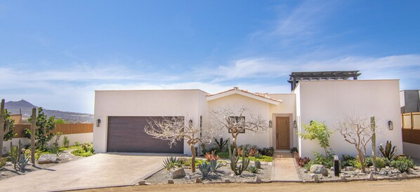 Una casa moderna de dos pisos con un garaje amplio, un jardín cuidado con diversos cactus y arbustos, y un cielo azul despejado.