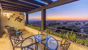 A covered patio with a glass table and chairs, a wall-mounted light fixture, and a view of the landscape during sunset. - Scenic Views Diamante 2Bdr Condo DRC #306 (Cabo San Lucas)