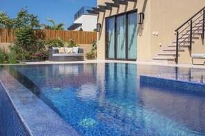 Ein Swimmingpool mit glatter, blauer Wasseroberfläche, ein Korbsessel mit Kissen und eine Holzlattenzaun.