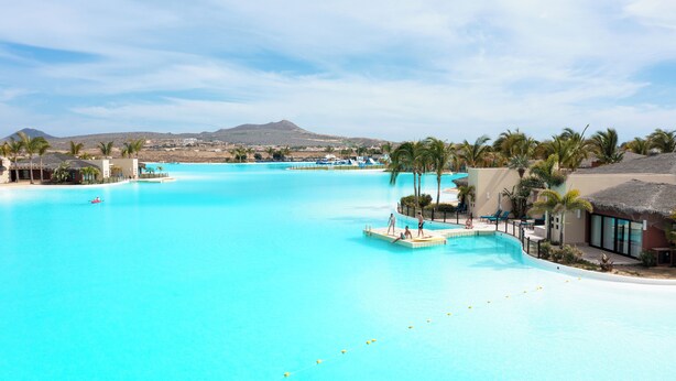 A large, clear blue swimming pool with a thatched-roof building and palm trees.