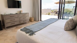 A bedroom with a large bed, a flat-screen TV mounted on the wall, a sliding glass door to the balcony, and a small wooden chest of drawers.