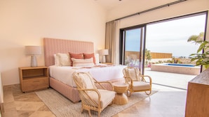 Habitación de hotel con una cama grande, dos mesitas de noche con lámparas, un rincón de estar con sillas de mimbre, una pequeña mesa redonda y un balcón con vista a la piscina.