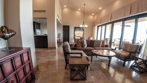 A spacious living area with a sofa, armchairs, and a coffee table. A kitchen is visible in the background, and there is a large window providing natural light.