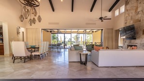 A spacious living area with a white sofa, a dining table with chairs, a TV mounted on the wall, and a view of the outdoors. - Luxurious Golf Villa #59 – 5-Bedroom (Cabo San Lucas)