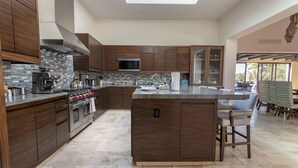 A modern kitchen with dark wood cabinets, a central island, stainless steel appliances, and a large window providing natural light. - Luxurious Golf Villa #59 – 5-Bedroom (Cabo San Lucas)
