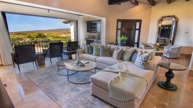 A spacious living room with a large sofa, coffee table, and chairs. There is a balcony with a view of the countryside.
