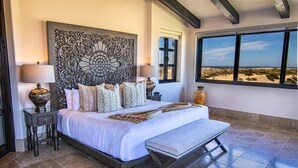 A bedroom with a large bed, two nightstands, a decorative headboard, a bench, and a view of the beach.