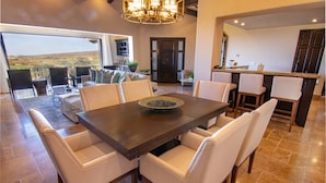 A spacious dining area with a large table and white chairs, a kitchen area with bar seating, and a view of the countryside through large windows.