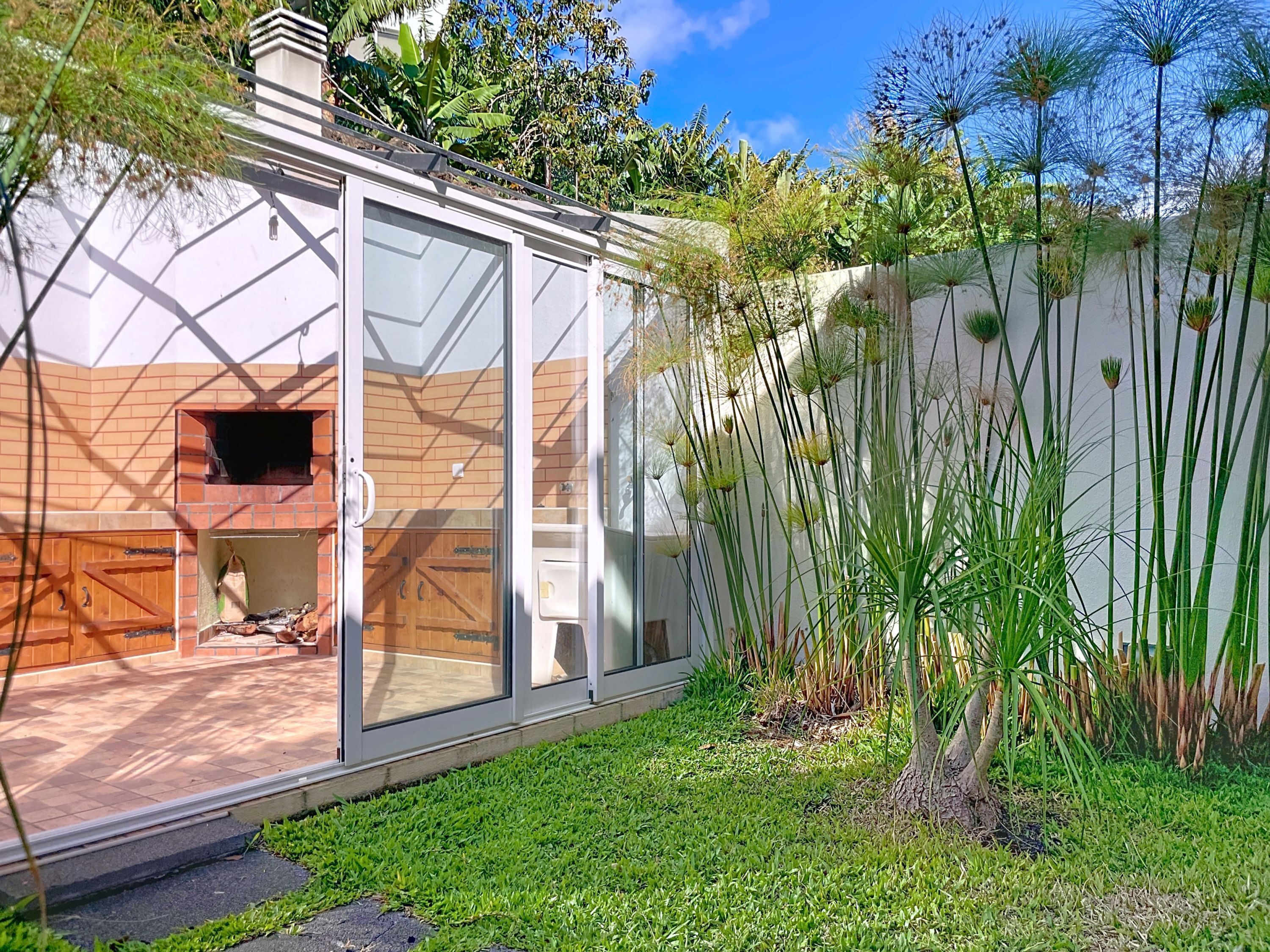 A modern house with a glass exterior, a brick chimney, and a well-maintained garden with tall grasses and a small tree.