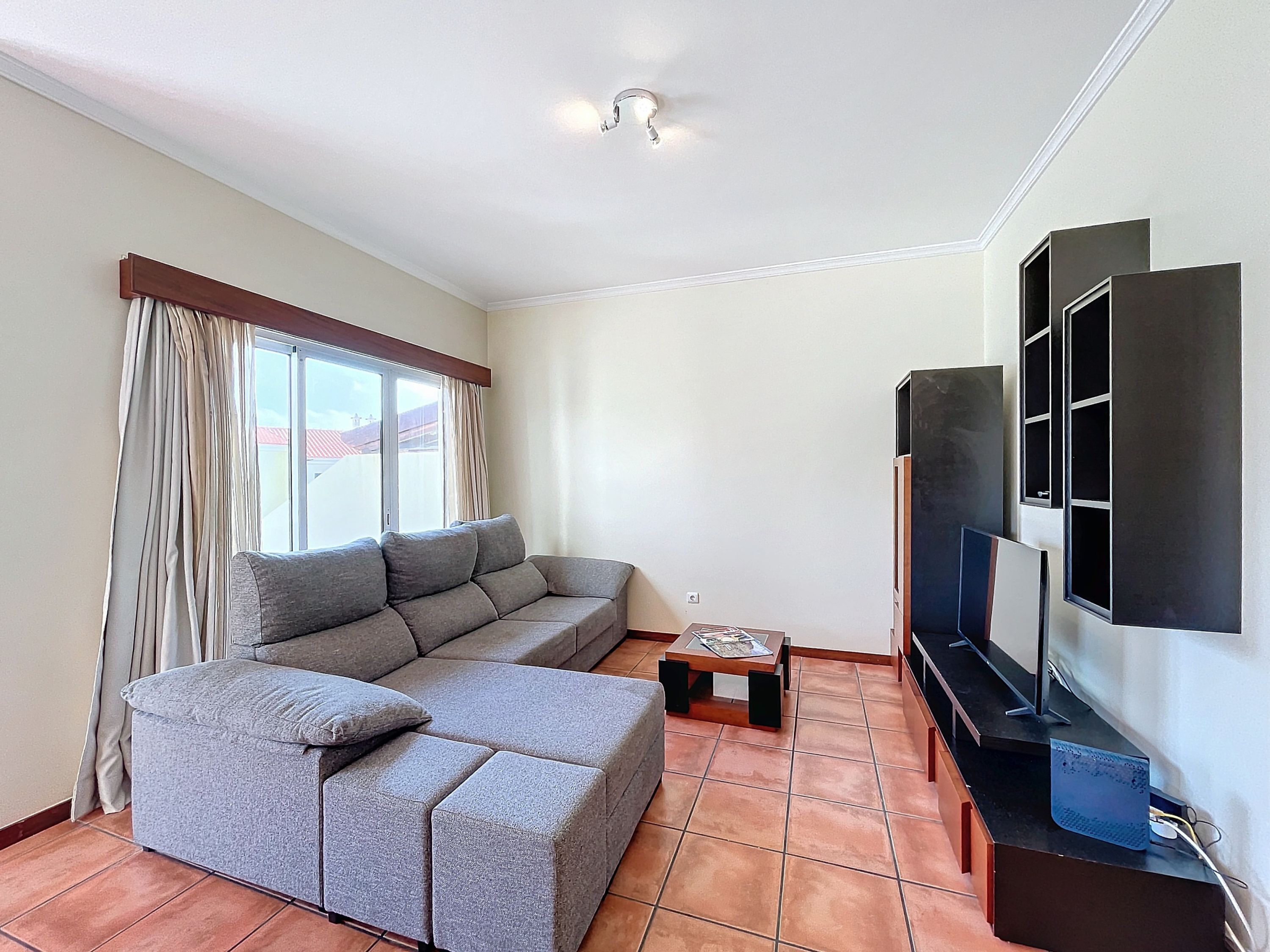 Una sala de estar con un sofá seccional gris, una mesa de centro, un televisor de pantalla plana montado en la pared y una ventana con cortinas.