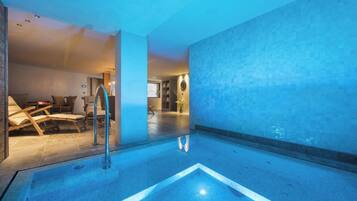 A modern indoor pool area with clear blue water, a lounge with chairs, and a visible staircase leading to another level.