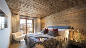A bedroom with a wooden ceiling, a large bed with multiple pillows, a bedside table with a lamp, and a chair. - Alpine Luxury Chalet Konk in Switzerland (Zermatt)