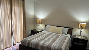 Un dormitorio con una cama, dos mesitas de noche con lámparas, una ventana con cortinas y una alfombra estampada.