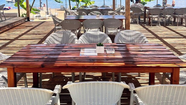 Un área de estar al aire libre con mesa de madera y sillas de mimbre blanco, con vista a una playa con sombrillas y gente.