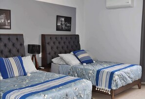 Una habitación de hotel con dos camas, cada una con cabecero capitonado, mesita de noche con lámpara y cuadros enmarcados en la pared.