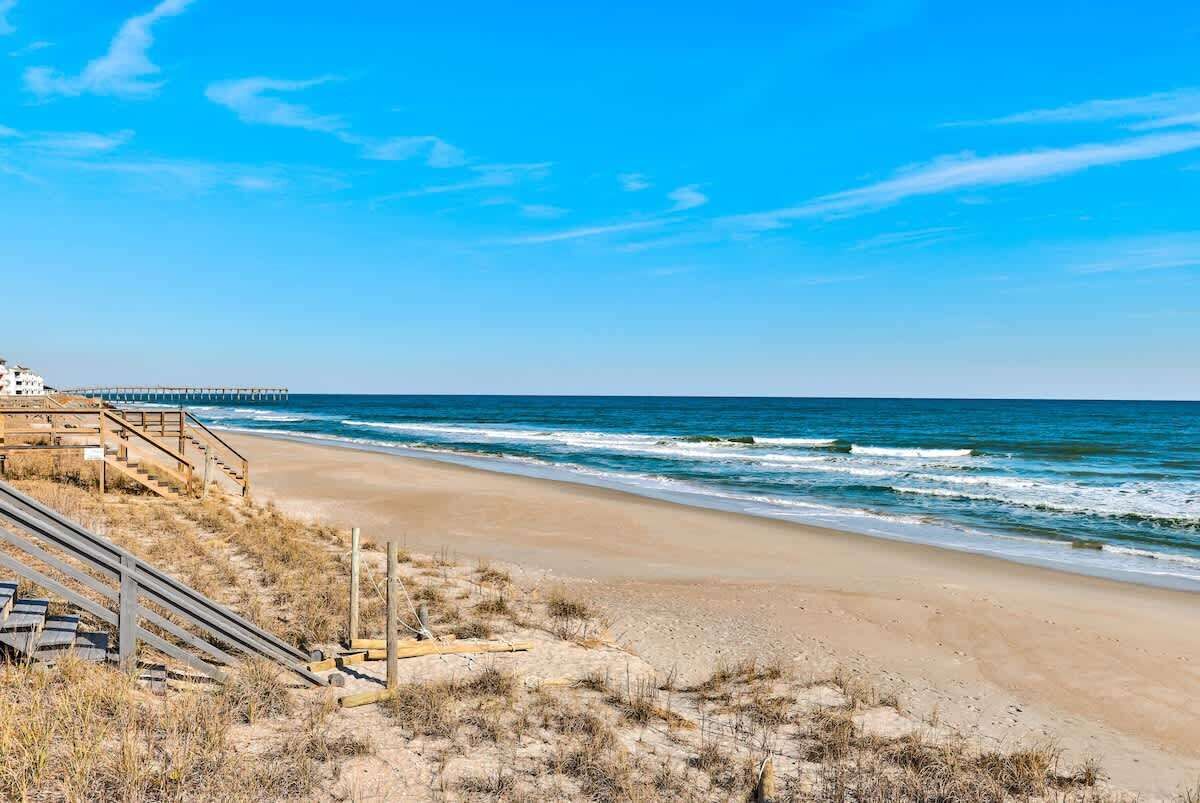 A beach with a wooden staircase leading down to the shore, a pier extending into the ocean, and a clear blue sky.
