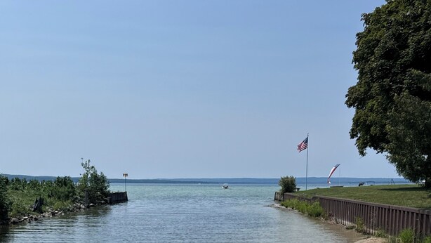 Ein Boot mit gepolsterten Sitzgelegenheiten und einem kleinen Tisch liegt an einem ruhigen Gewässer mit Uferwiese und Fahnenmast.