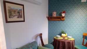A cozy living room with a teal sofa, a wooden chair, a small round table with a brown tablecloth, and a wall-mounted shelf with decorative items. - MAISONETTE FOR 2 PEOPLE NEAR EUROPA PARK AND RULANTICA  (BINDERNHEIM)