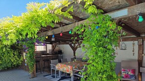 A covered outdoor seating area with a table and chairs, surrounded by greenery and a wooden structure. - MAISONETTE FOR 2 PEOPLE NEAR EUROPA PARK AND RULANTICA  (BINDERNHEIM)