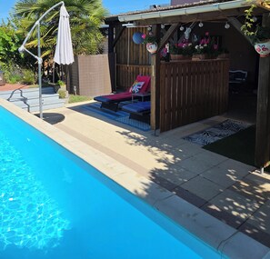 A swimming pool with a wooden deck area, a covered seating area with cushions, and hanging flower pots.