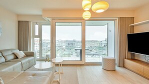 Un salon moderne avec un canapé, une table basse, un téléviseur et une grande fenêtre donnant sur la ville.
