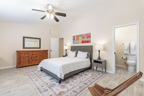 Een slaapkamer met een bed, nachtkastjes, een plafondventilator, een dressoir, een vloerkleed en een zichtbare badkamer door een geopende deur.