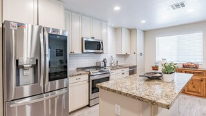 A modern kitchen with stainless steel appliances, white cabinetry, granite countertops, and a window with blinds. - Gilbert AZ 4BR Vacation Home Remodeled Garage (Gilbert)
