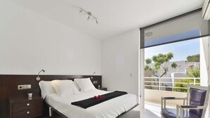 A modern bedroom with a large bed, a nightstand, a chair, and a balcony with a view of the city.