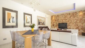 A modern living room with a stone accent wall, a flat-screen TV, and a dining area with a wooden table and wireframe chairs.