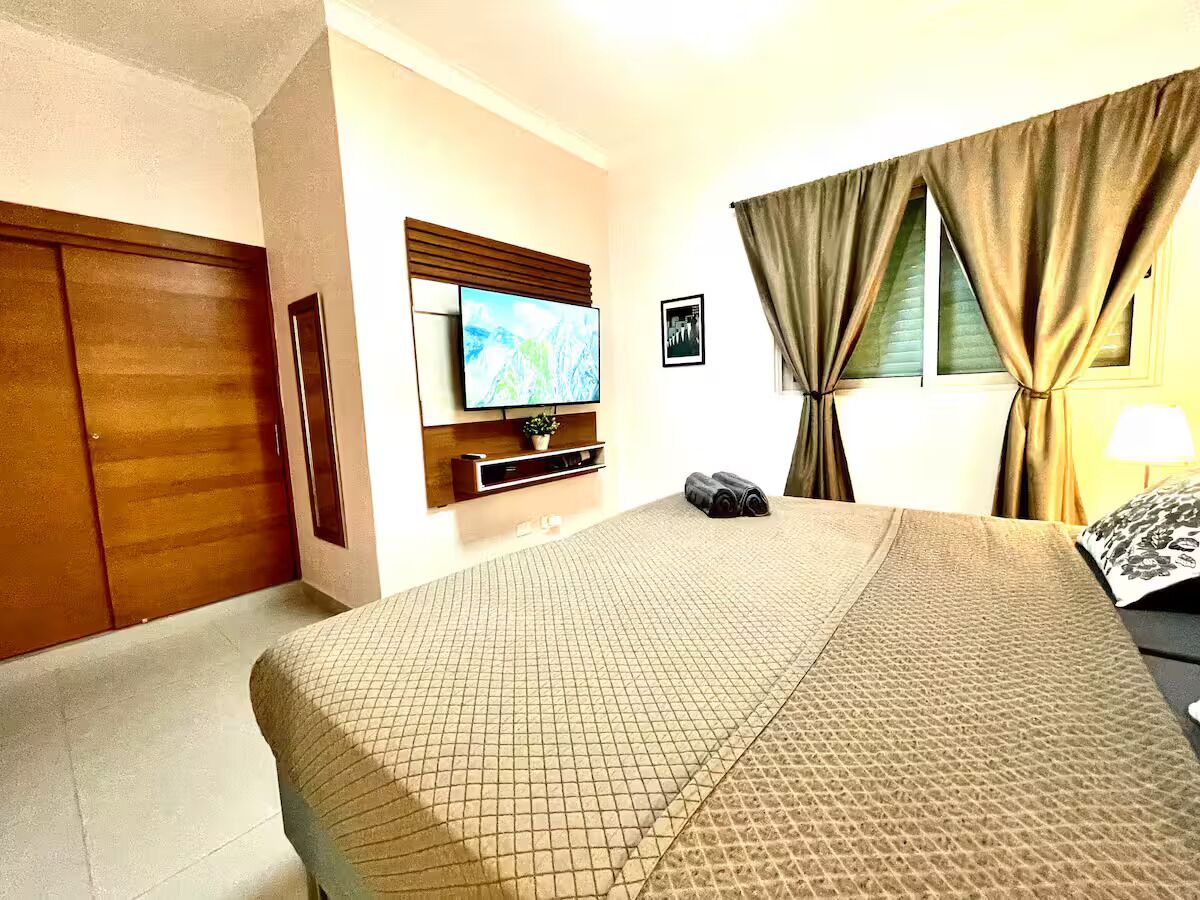 Une chambre d’hôtel avec un lit, un téléviseur fixé au mur, une fenêtre avec des rideaux et une armoire en bois.