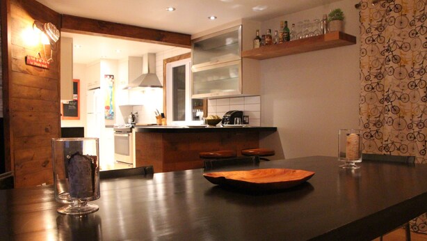 A modern kitchen with a dark countertop, a wooden bar area, and a patterned wallpaper. - Cozy Chalet Near Mont-Sainte-Anne | Sleeps 8–10 | Fireplace | EV Charger (Saint-Ferreol-les-Neiges)