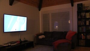 A living room with a television, a sofa, a bookshelf, and a window. - Cozy Chalet Near Mont-Sainte-Anne | Sleeps 8–10 | Fireplace | EV Charger (Saint-Ferreol-les-Neiges)