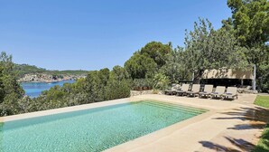 A rectangular swimming pool with clear blue water, surrounded by a stone deck. There are lounge chairs and a covered seating area with a view of the sea and hills in the background. - Amazing Villa With Swimming Pool (San Juan de Alicante)