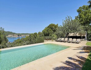 A rectangular swimming pool with clear blue water, surrounded by a stone deck. There are lounge chairs and a covered seating area with a view of the sea and hills in the background. - Amazing Villa With Swimming Pool (San Juan de Alicante)