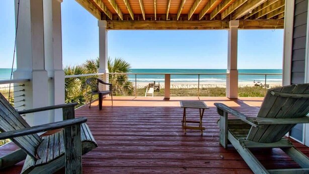 A wooden deck with two lounge chairs and a small table, overlooking a beach with a clear view of the ocean. - One Step Away - Sleeps 28 (Carolina Beach)