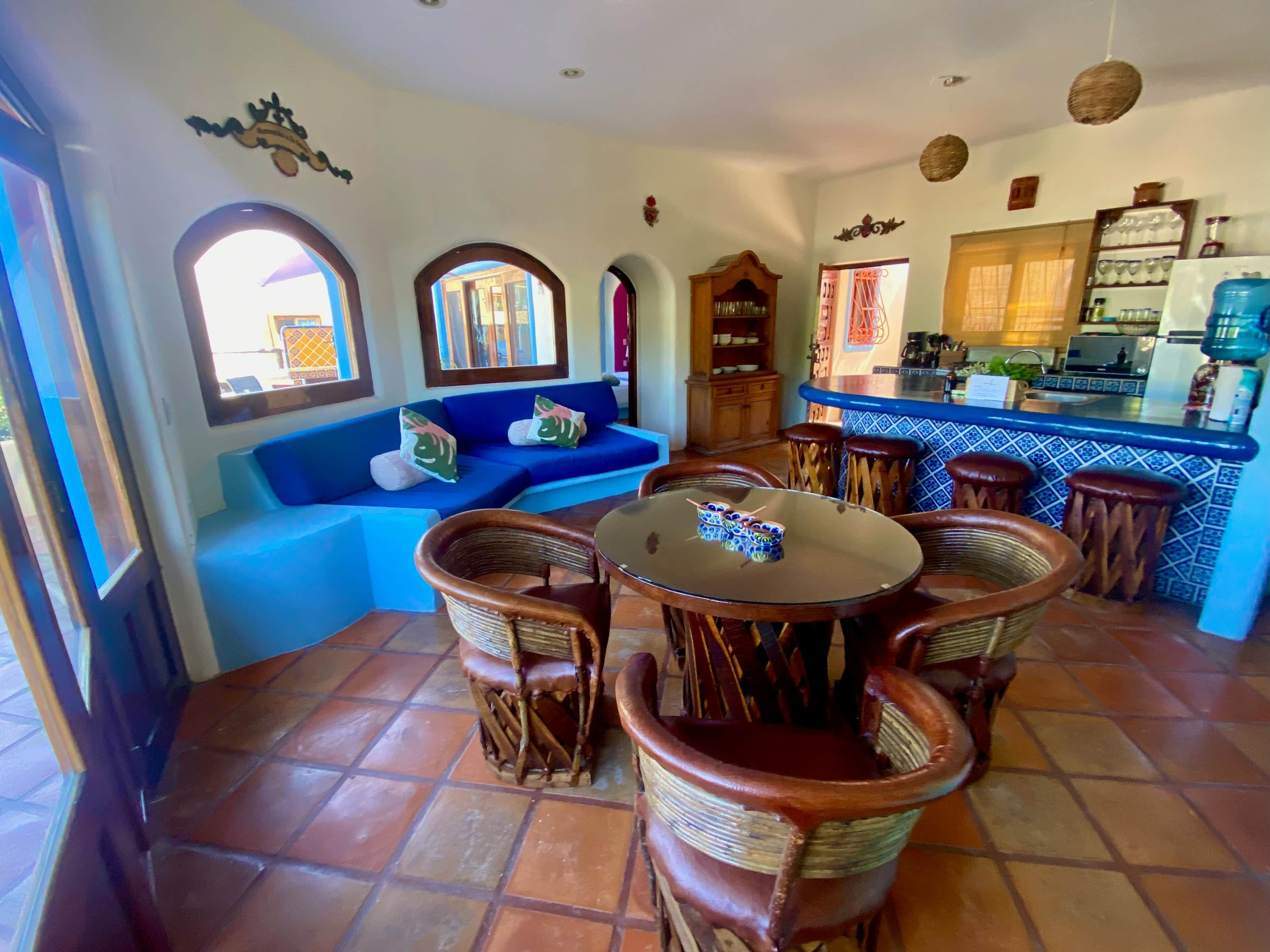 Un área de estar acogedora con un sofá azul, una mesa redonda de vidrio, sillas de mimbre y una cocina al fondo.