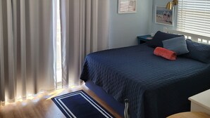 Une chambre à coucher avec un lit, des rideaux, un tableau mural, des photos encadrées et une fenêtre avec des stores.