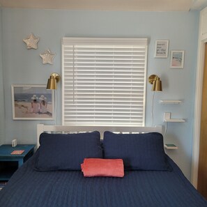 Quarto com cama, janela com persianas, mesa de cabeceira com abajur e decoração de parede incluindo estrelas e um porta-retrato.