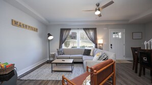 A living room with a sofa, coffee table, and a chair. There is a ceiling fan, a window with curtains, and a door.