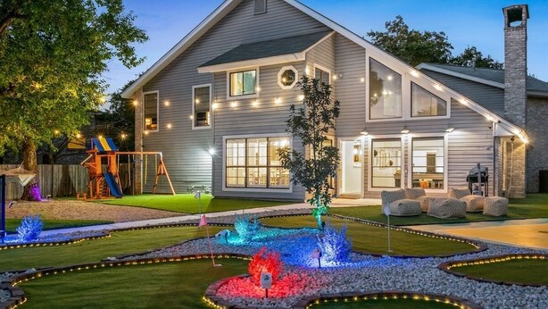 A two-story house with a well-maintained lawn, a swing set, and a decorative garden with illuminated plants. - The Ultimate Getaway: Pool, Pickleball, Mini Golf (San Antonio)